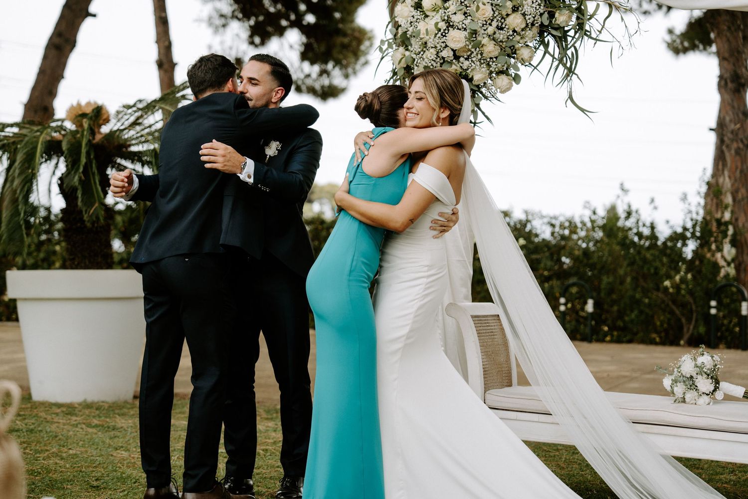 Bride and groom embracing family members after outdoor wedding ceremony near floral arch.