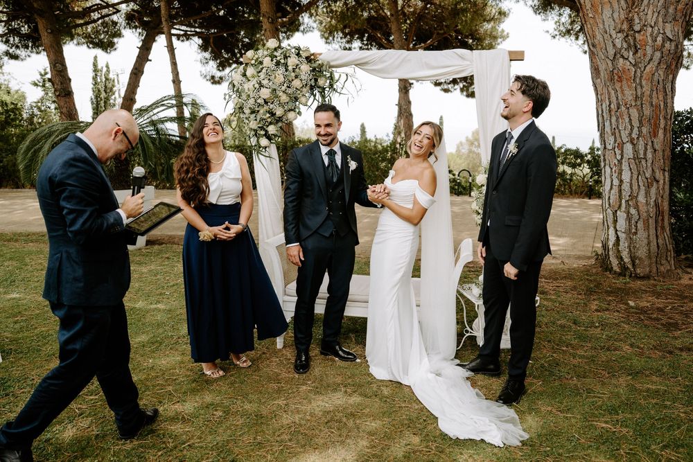 Bride and groom laughing with wedding party members at outdoor ceremony beneath pine trees.
