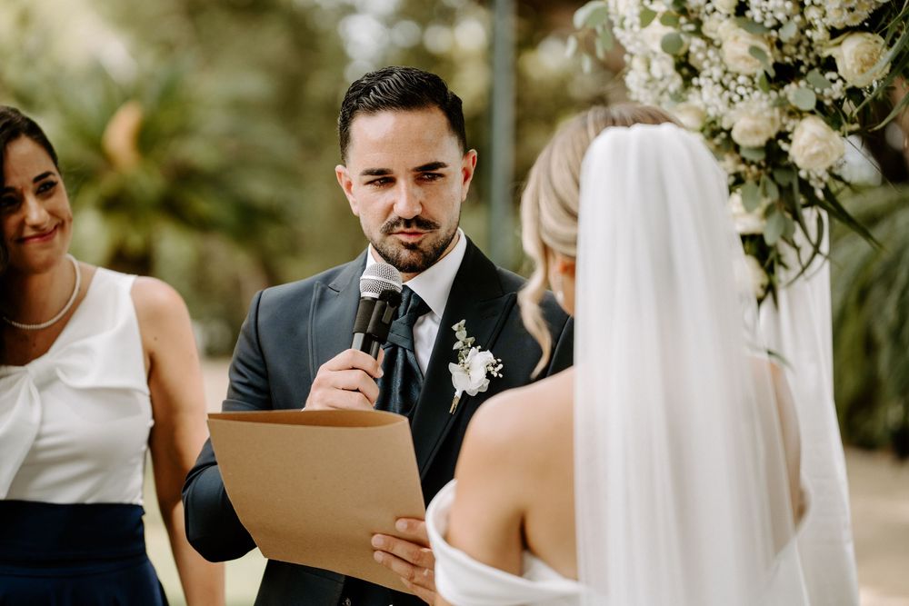 Groom in dark suit reads heartfelt vows to bride during elegant outdoor wedding ceremony.