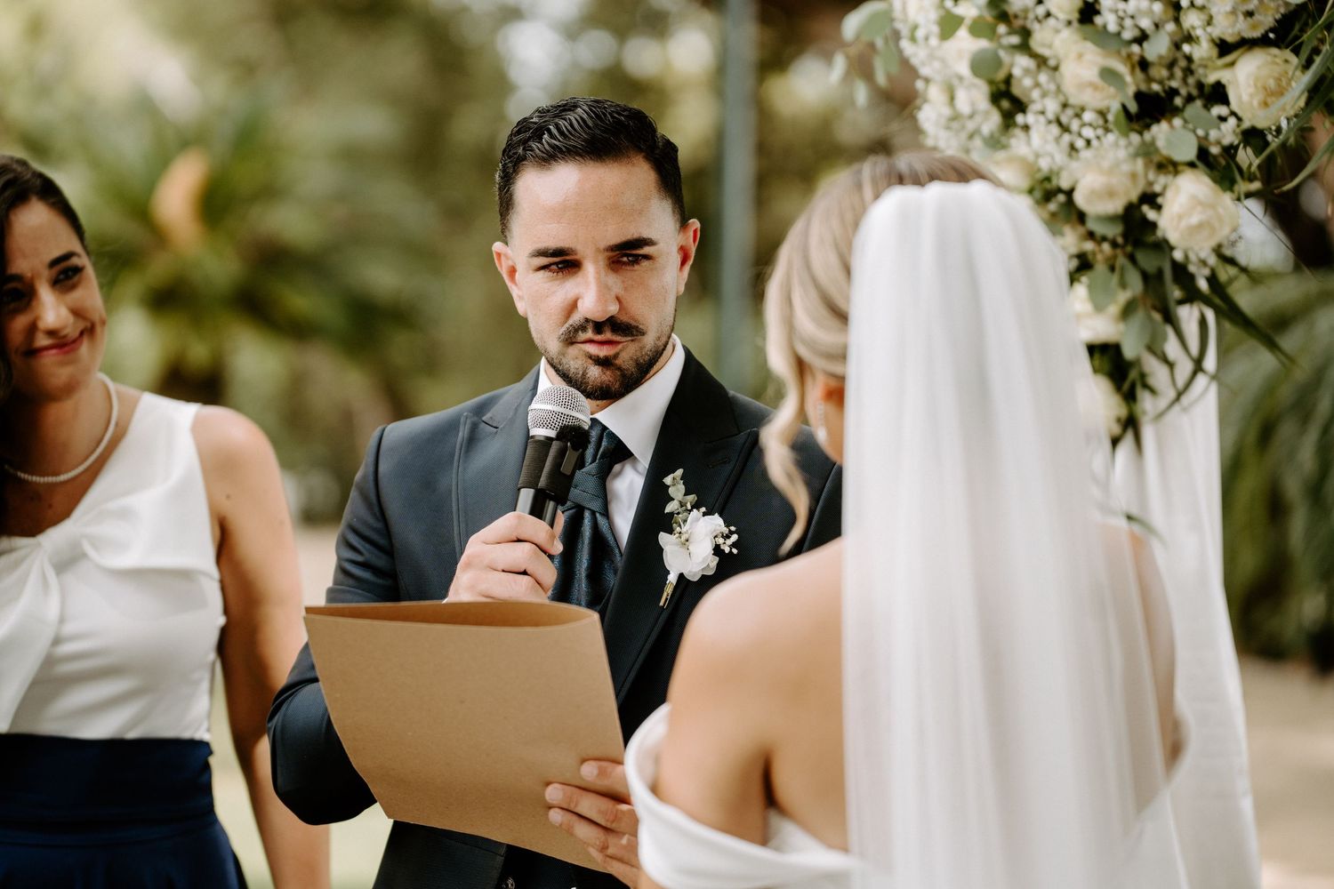 Groom in dark suit reads heartfelt vows to bride during elegant outdoor wedding ceremony.