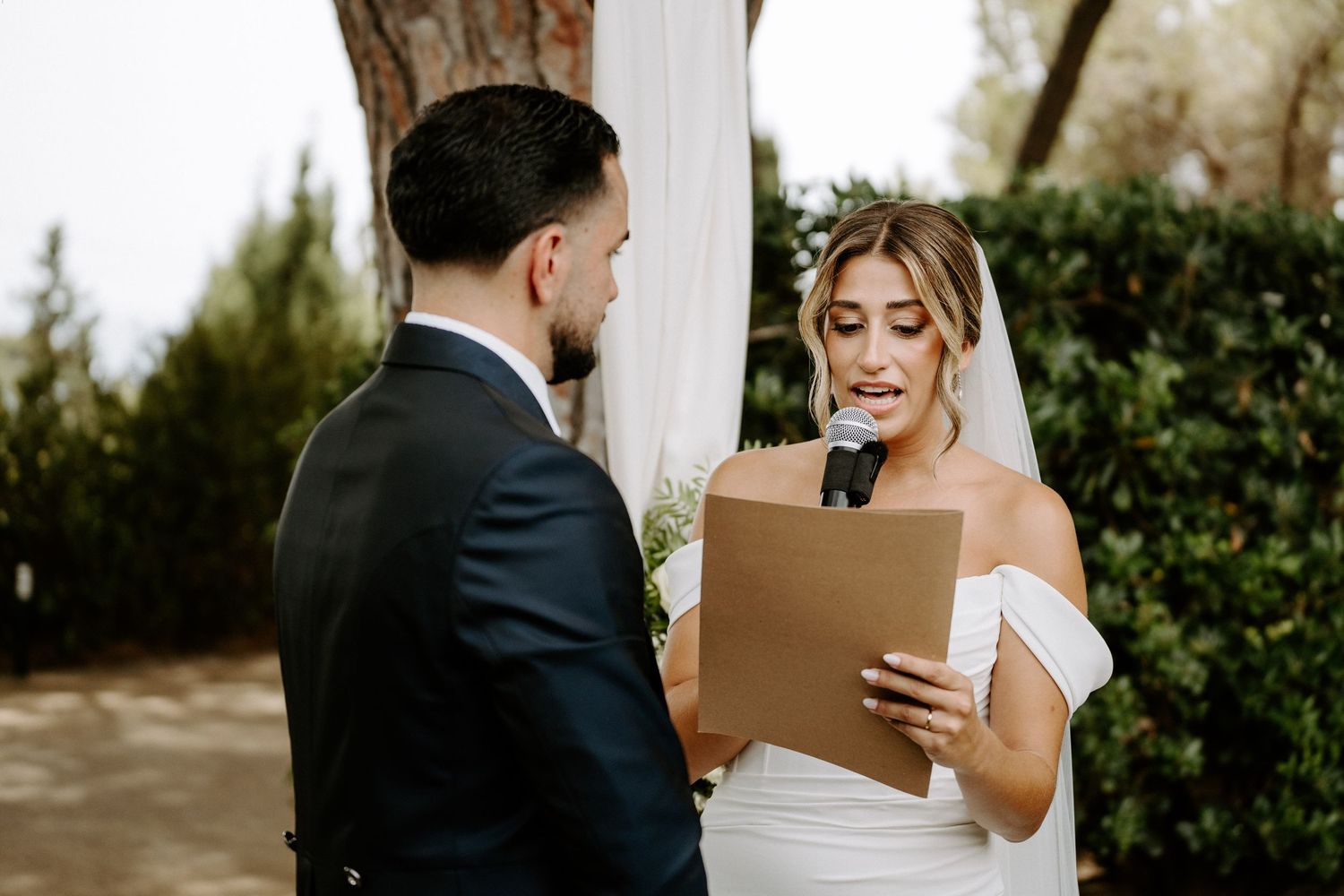 Bride in off-shoulder gown reads vows from paper at outdoor wedding ceremony with groom in black suit.