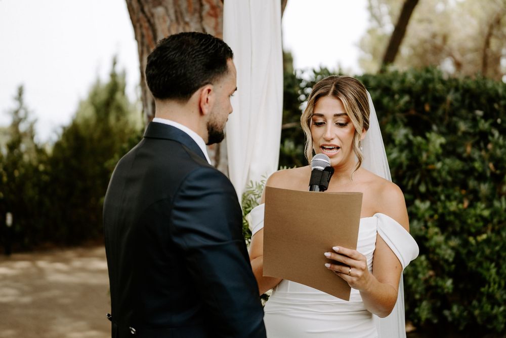 Bride in off-shoulder gown reads vows from paper at outdoor wedding ceremony with groom in black suit.