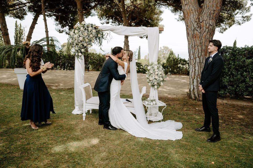 Couple shares first kiss under floral arch at outdoor wedding ceremony with guests and officiant nearby.