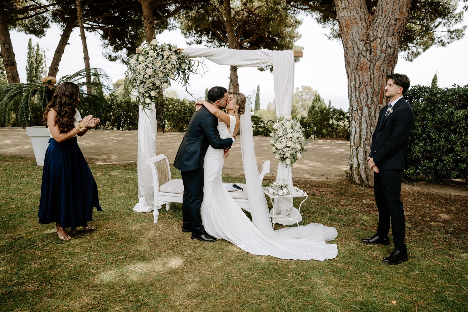 Couple shares first kiss under floral arch at outdoor wedding ceremony with guests and officiant nearby.