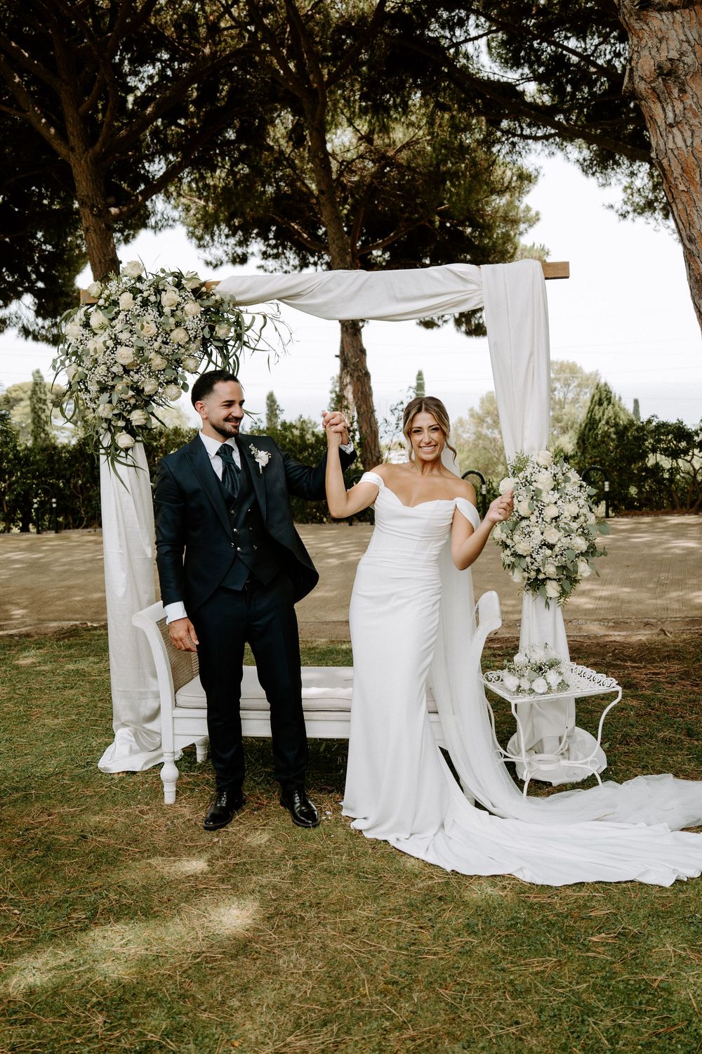 Joyful bride and groom celebrate under floral arch with champagne at outdoor garden wedding.
