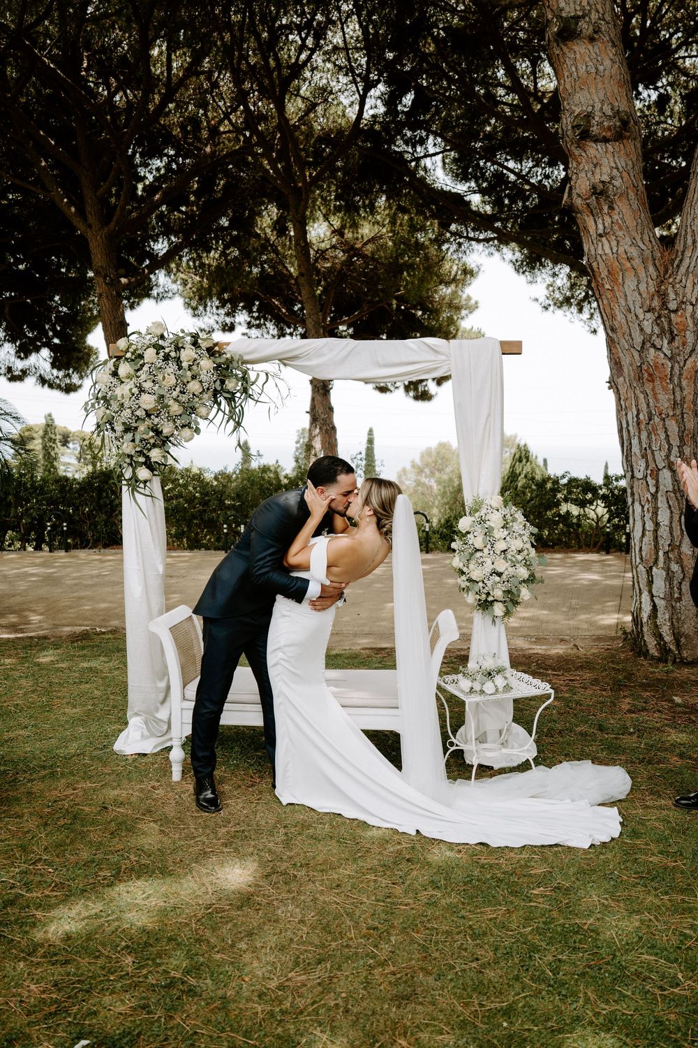 Newlyweds share first kiss under white draped arch decorated with white floral arrangements outdoors.