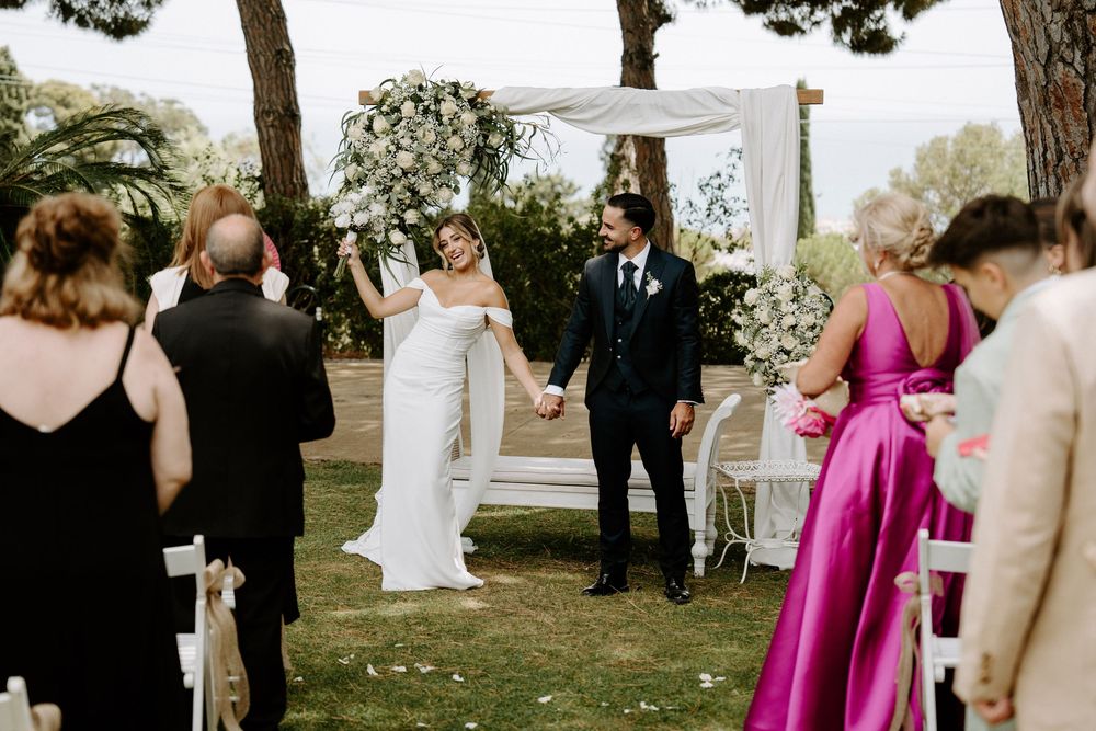 Bride and groom holding hands at outdoor wedding ceremony with floral arch, guests seated on white chairs.