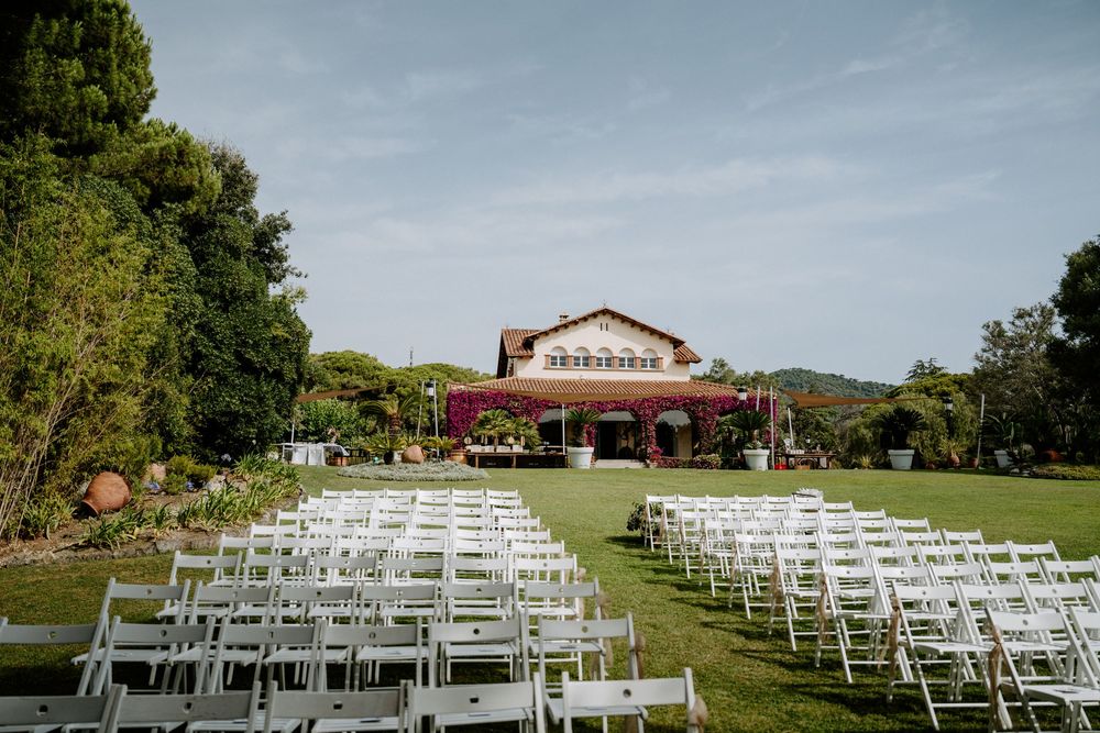 Outdoor wedding ceremony setup with white chairs on green lawn facing a rustic venue adorned with purple flowers and lush trees.