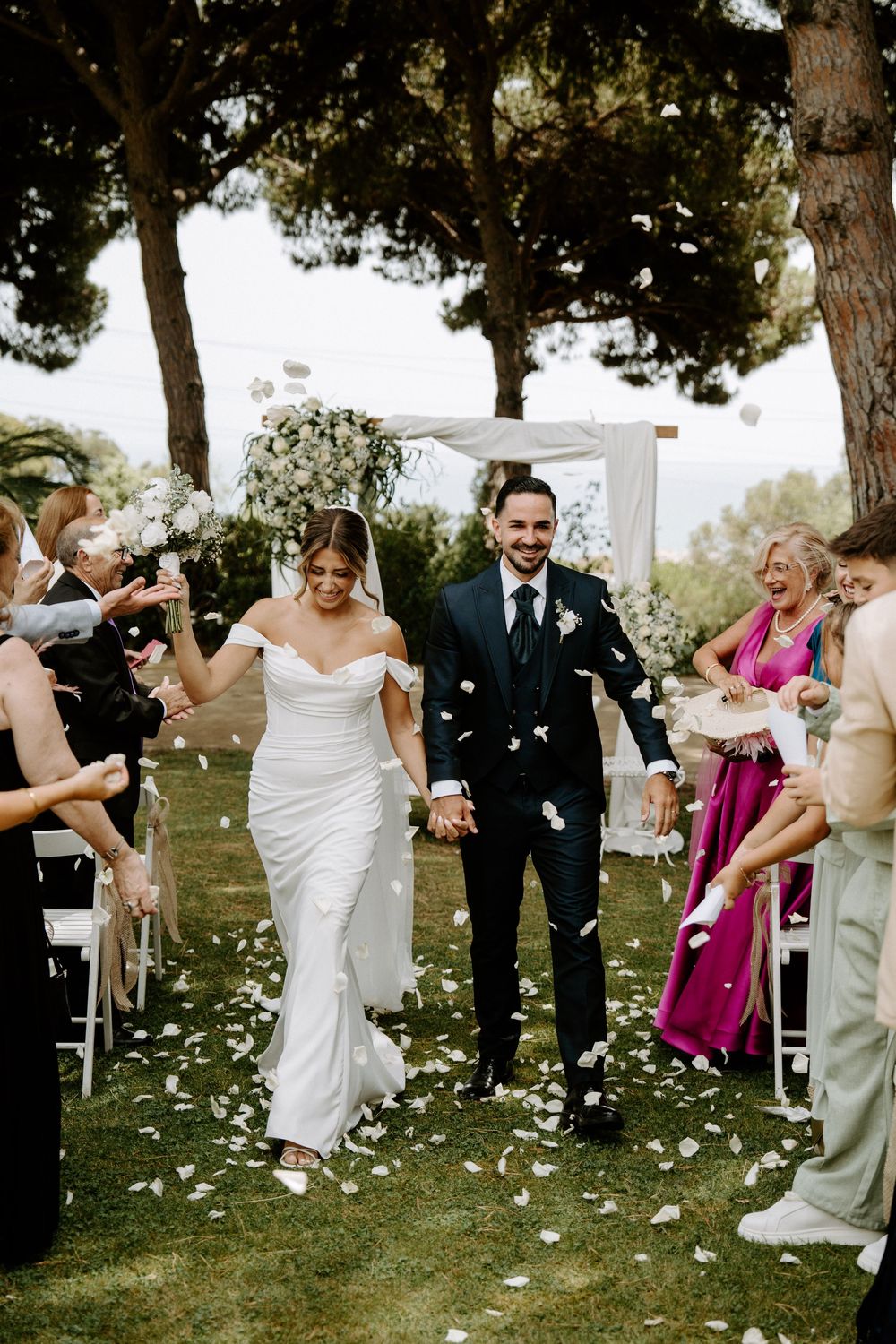 Newlyweds walk down aisle as guests shower them with white rose petals at outdoor ceremony.