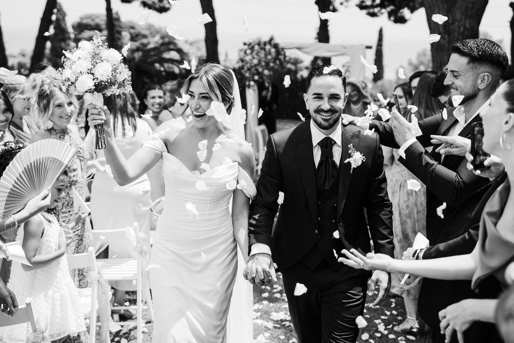 Black and white photo of smiling newlyweds walking through petal toss after outdoor wedding ceremony.