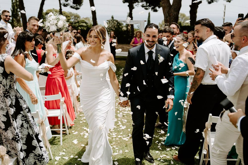 Beaming newlyweds walk through confetti shower as guests celebrate at elegant outdoor wedding ceremony.