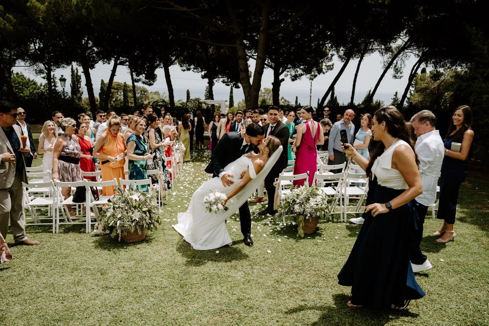 Bride and groom share romantic dip kiss while walking down aisle surrounded by cheering wedding guests outdoors.