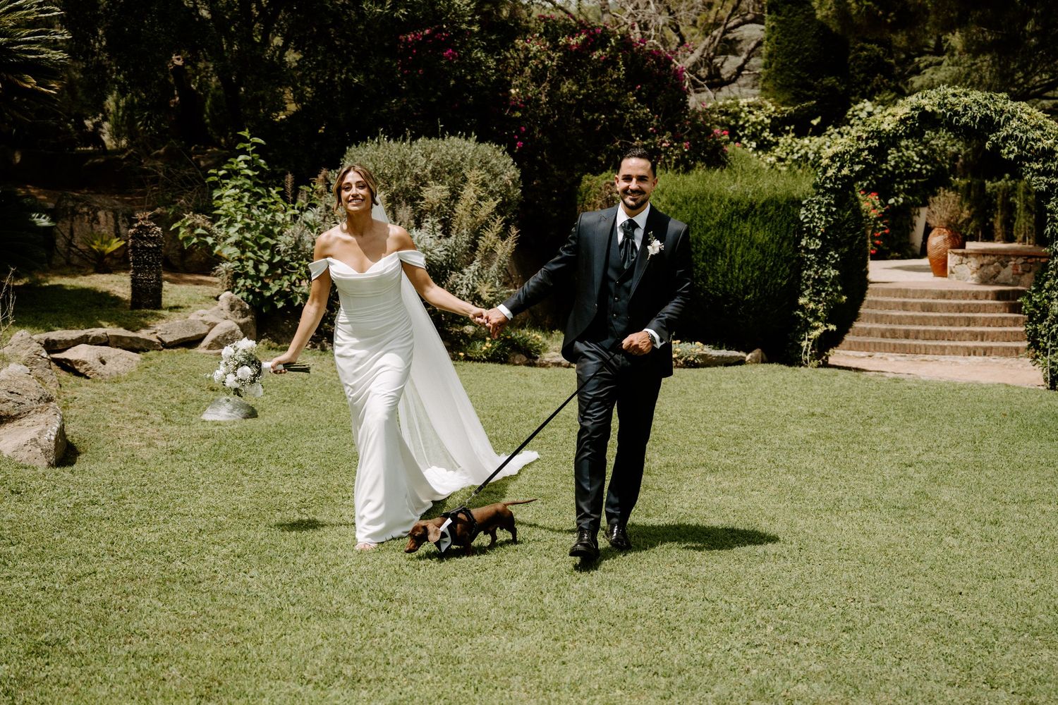 Happy bride and groom walk their small dog across lush green garden at wedding venue.