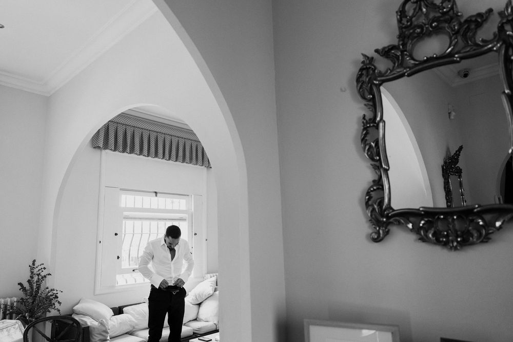 Black and white photo of a groom adjusting his shirt in an elegant room with an ornate mirror and arched doorway.