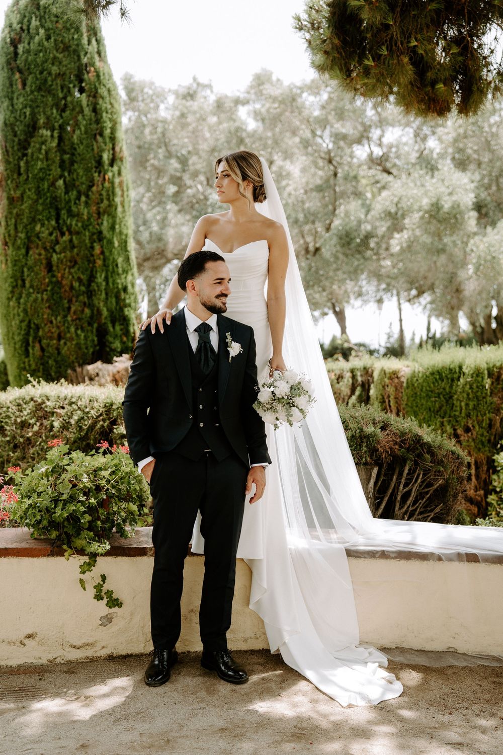 Bride and groom posing in lush garden, bride in strapless gown with veil, groom in black suit.
