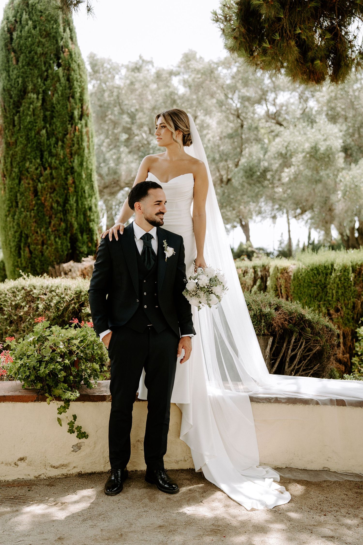 Bride and groom posing in lush garden, bride in strapless gown with veil, groom in black suit.