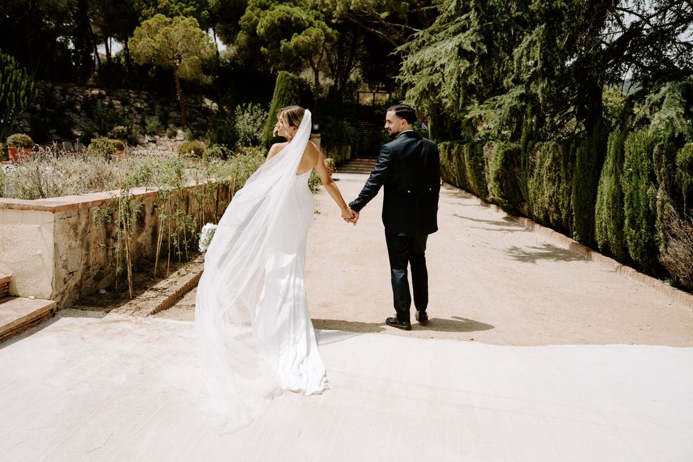 Bride and groom holding hands walking along a garden path, her long veil trailing behind in a lush outdoor wedding setting.