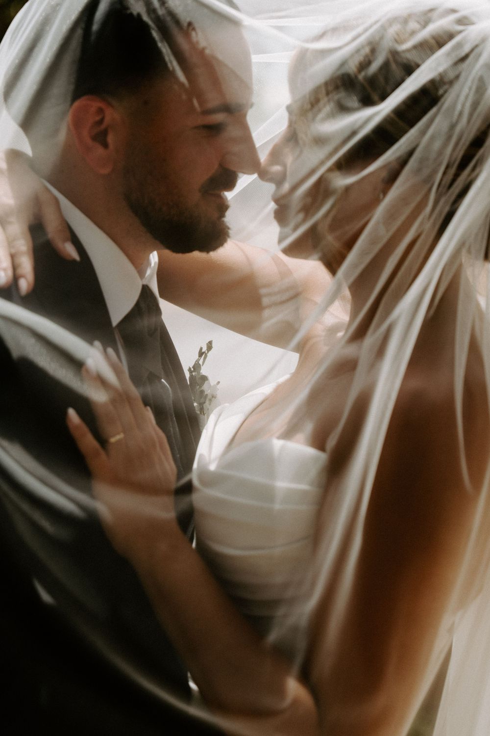 Bride and groom share an intimate moment under a flowing wedding veil, smiling at each other in soft romantic light.