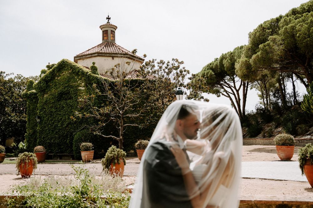 Bride and groom kissing under a veil at a romantic Italian villa wedding with lush gardens and a domed chapel.