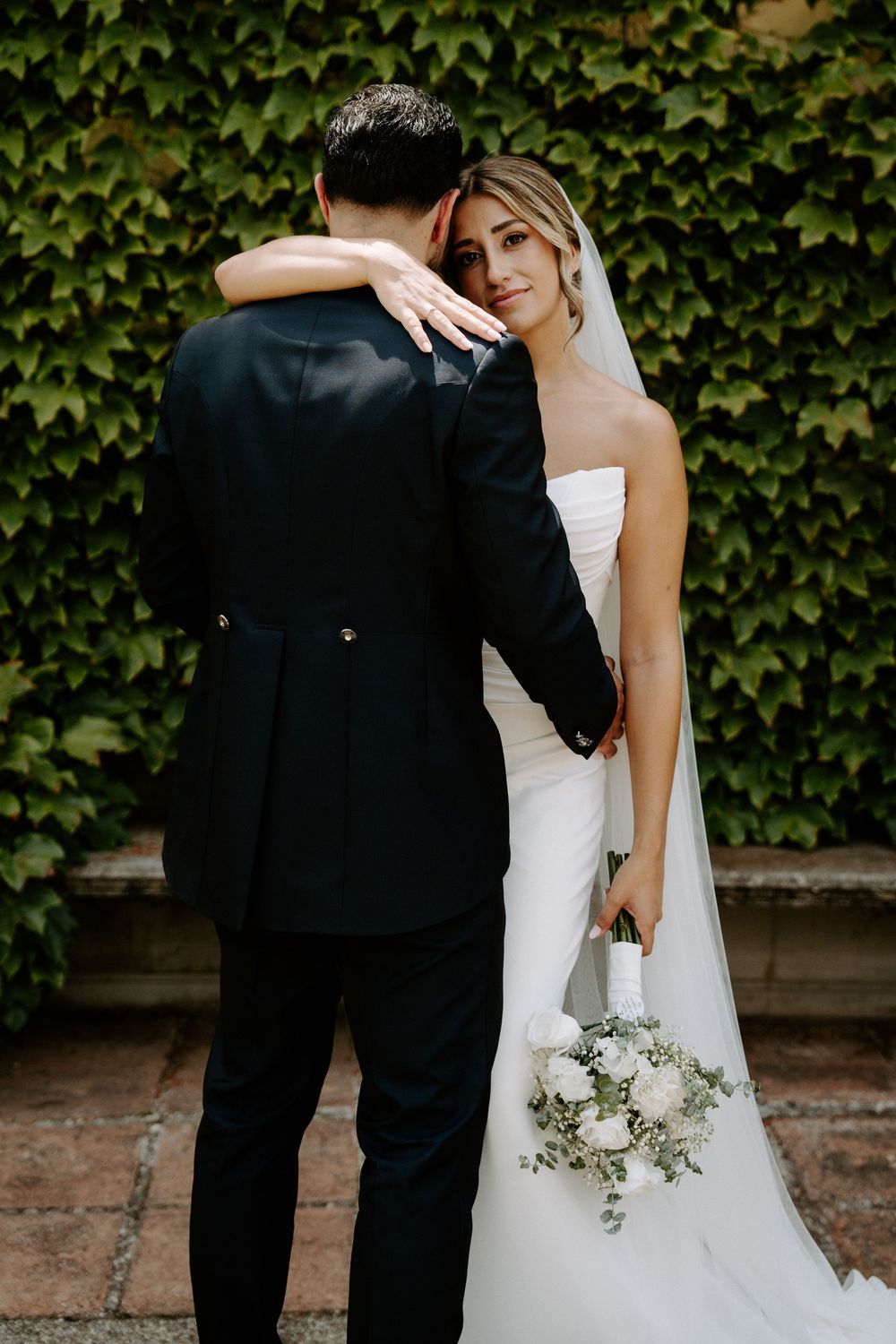 Bride in white strapless gown holds bouquet, embracing groom in black suit before ivy-covered wall.