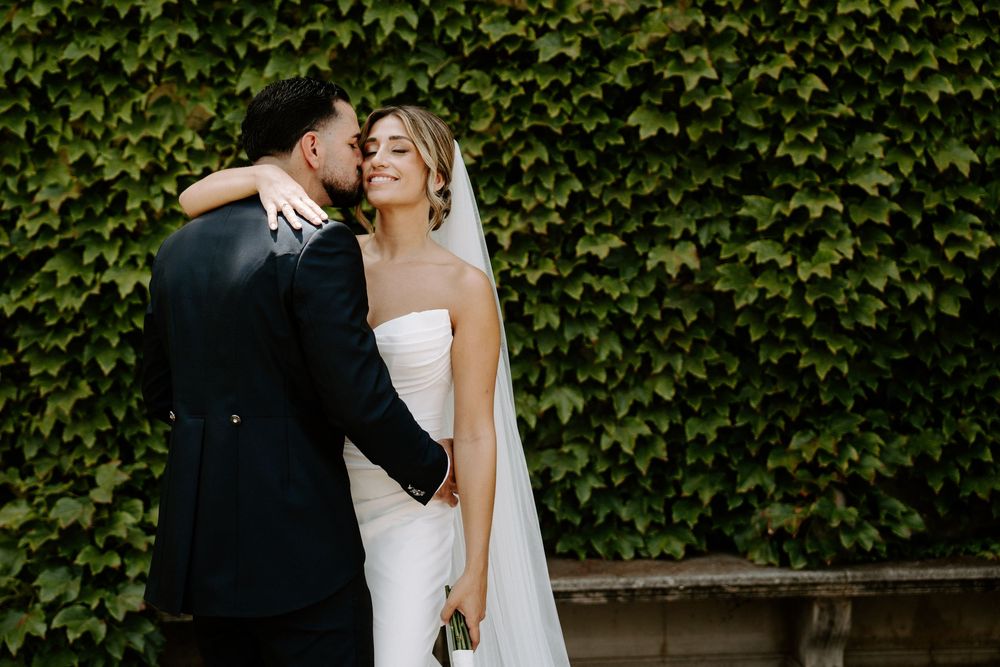 Bride in white strapless gown smiles as groom in black suit kisses her cheek before ivy-covered wall.