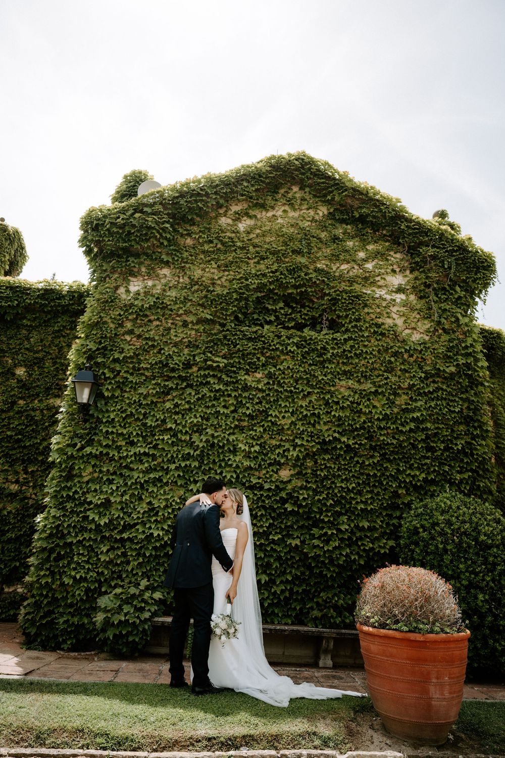 Bride and groom kissing in front of an ivy-covered stone wall with a terracotta pot in a lush garden.