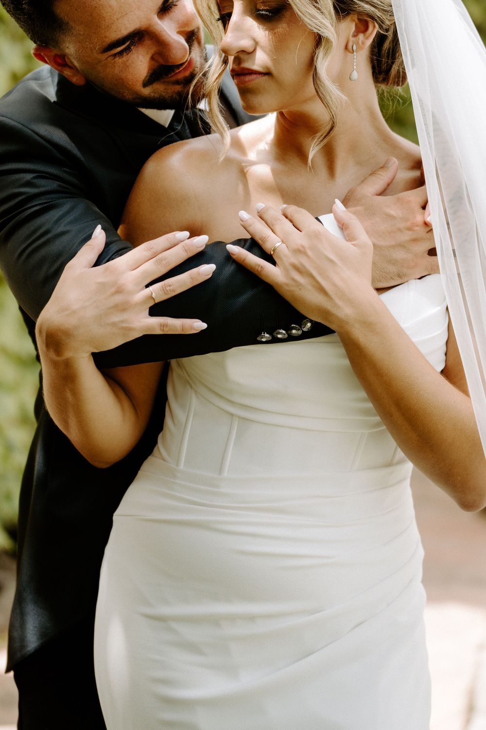 Groom in black suit embracing bride in white strapless wedding dress and veil, showing wedding rings.