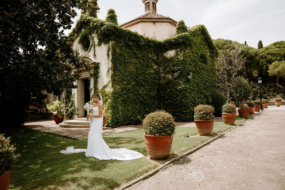 Bride in white gown with train stands before ivy-covered villa surrounded by terracotta pots.