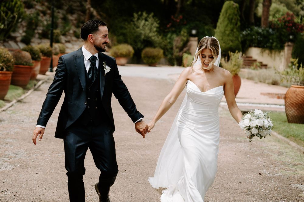 Bride and groom walking hand in hand outdoors, smiling, bride holding white bouquet in elegant strapless gown.