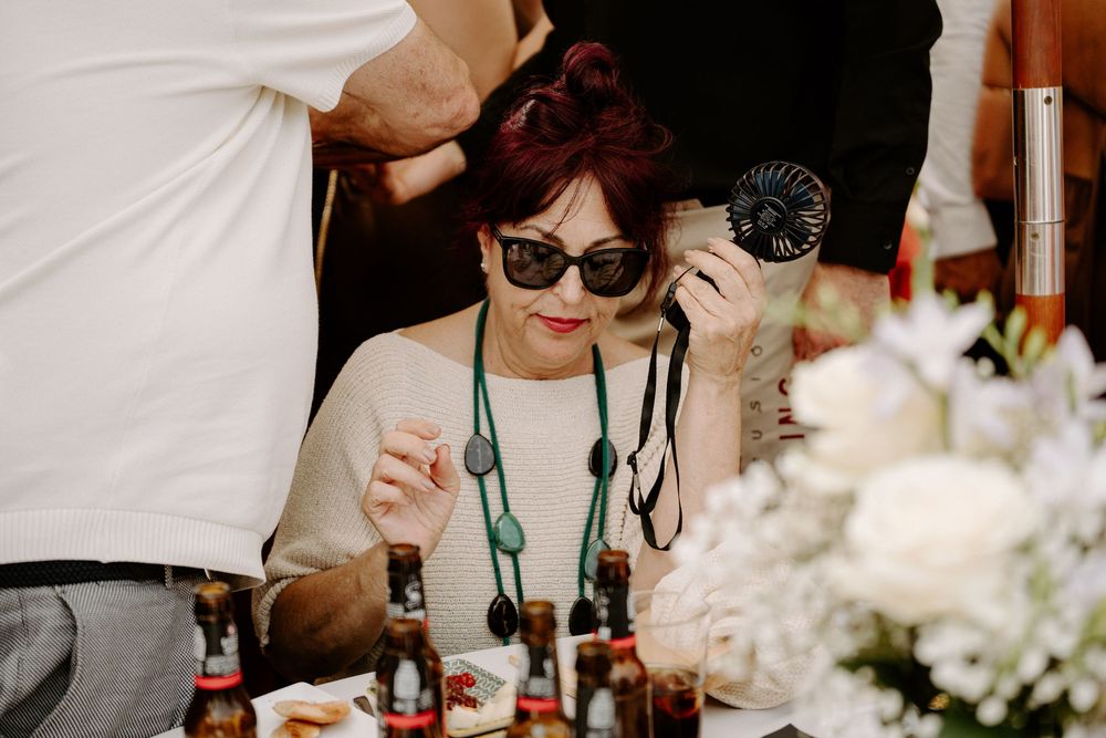 Woman with red hair and sunglasses holding a fan at an outdoor event with bottles and white flowers.