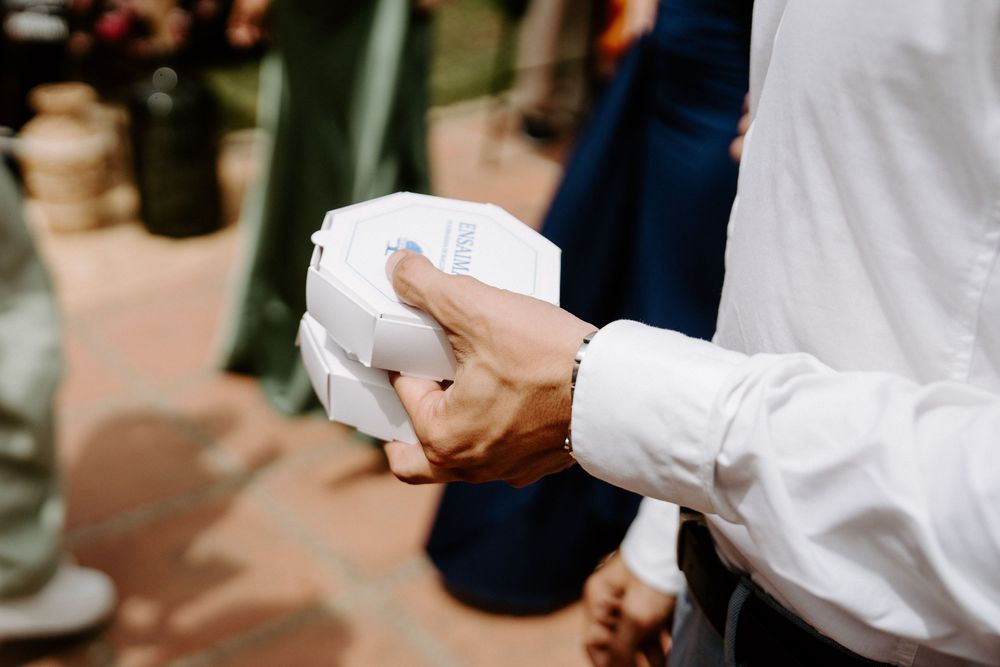 Close-up of a man in white shirt holding a small white gift box at an outdoor wedding.