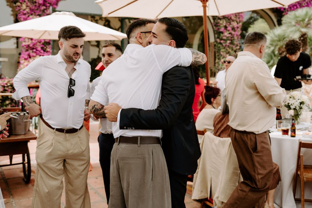 Two men share a warm embrace at an outdoor wedding reception surrounded by guests under white umbrellas.