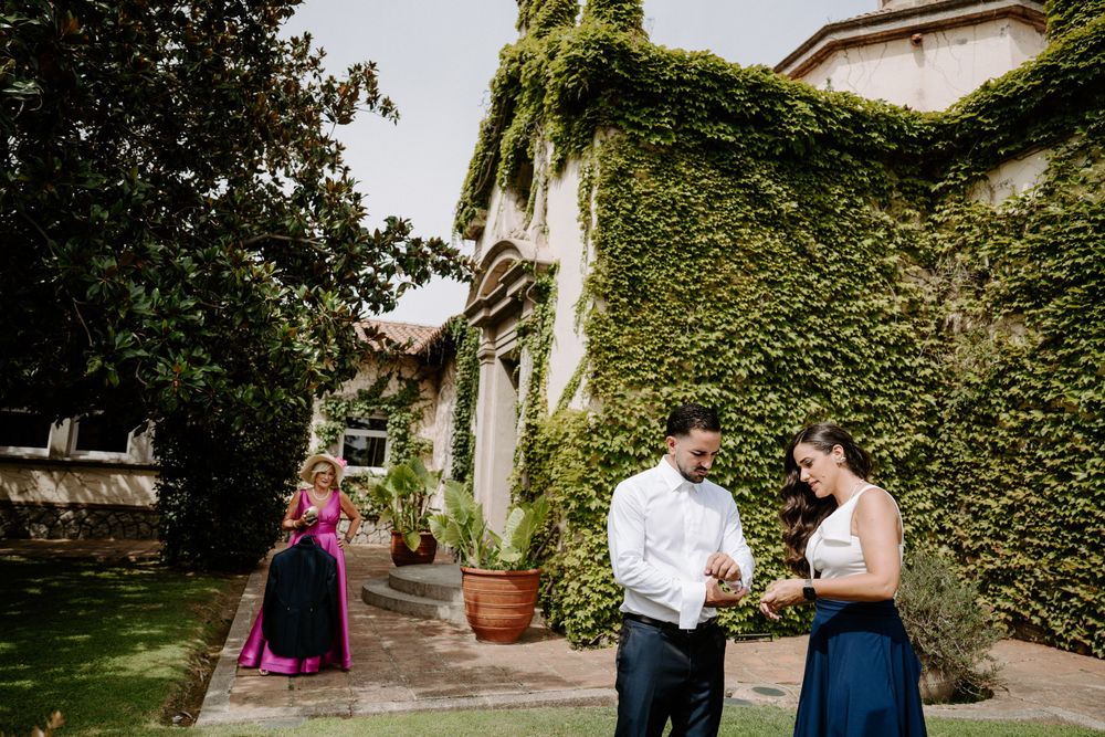 Couple exchanging vows outdoors at ivy-covered villa wedding venue with bridesmaid in blue dress nearby.