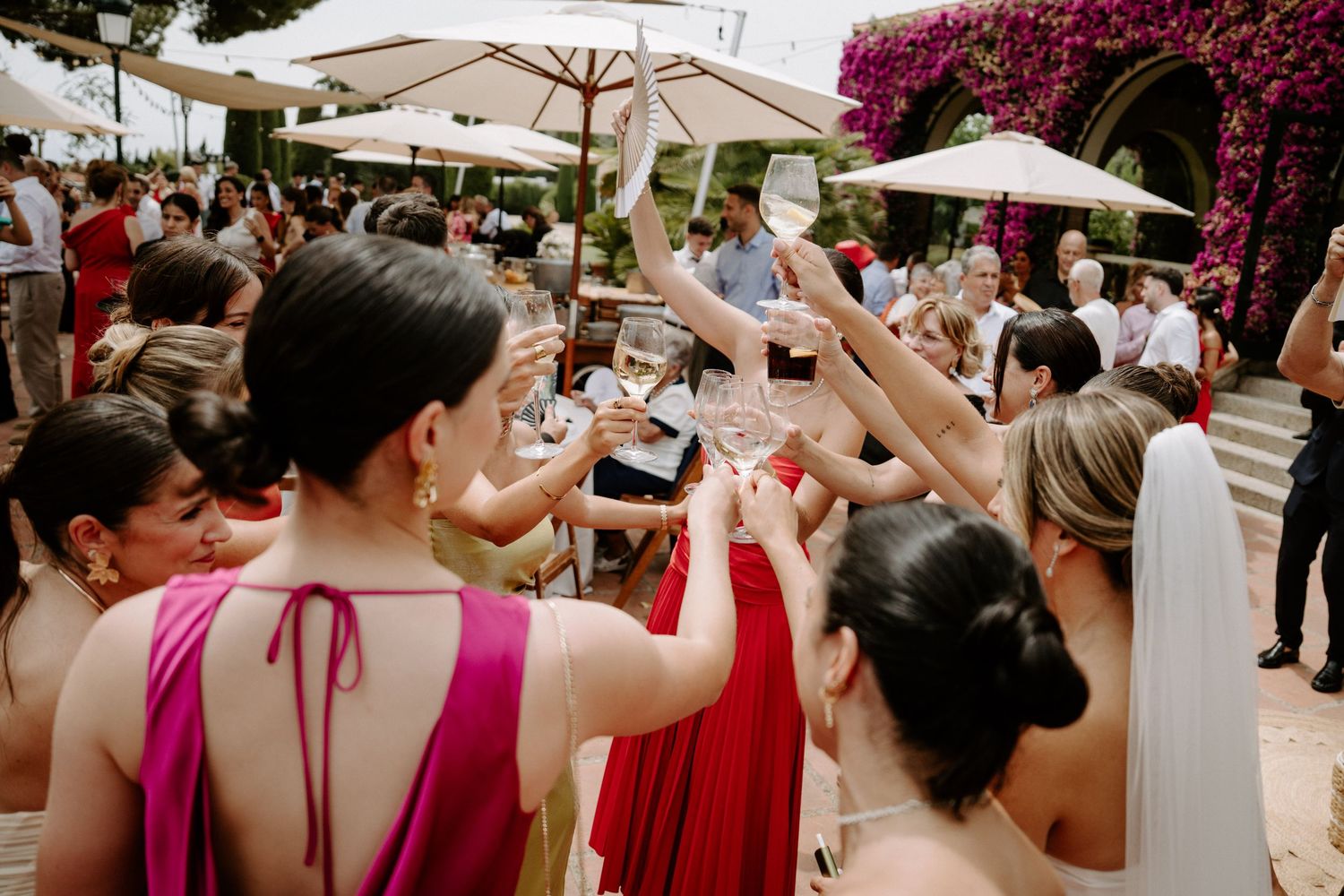 Guests raising champagne glasses in a toast at an outdoor wedding reception with white umbrellas and pink flowers.
