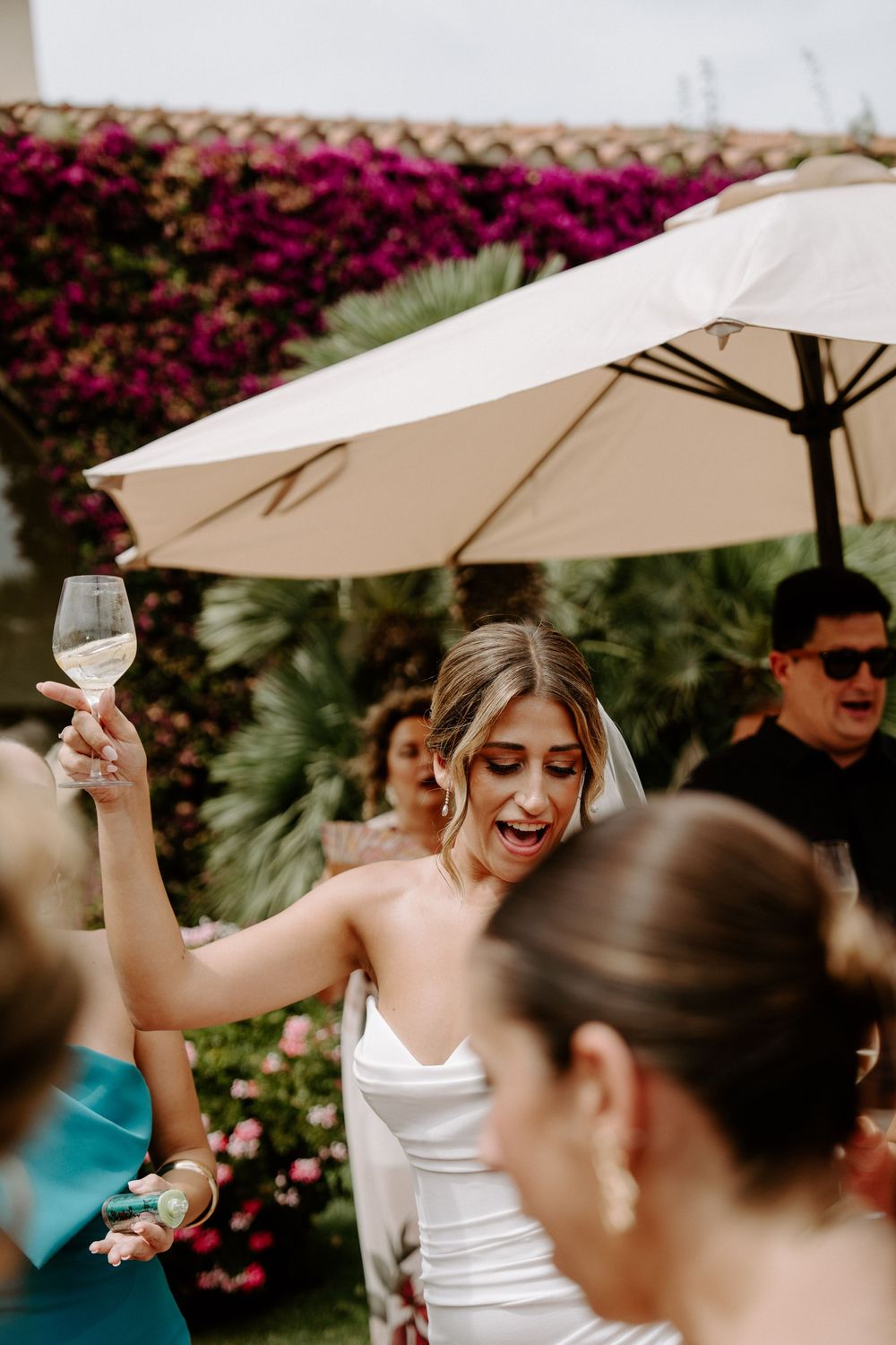 Bride in white dress raises champagne glass celebrating outdoors at wedding reception with floral backdrop.