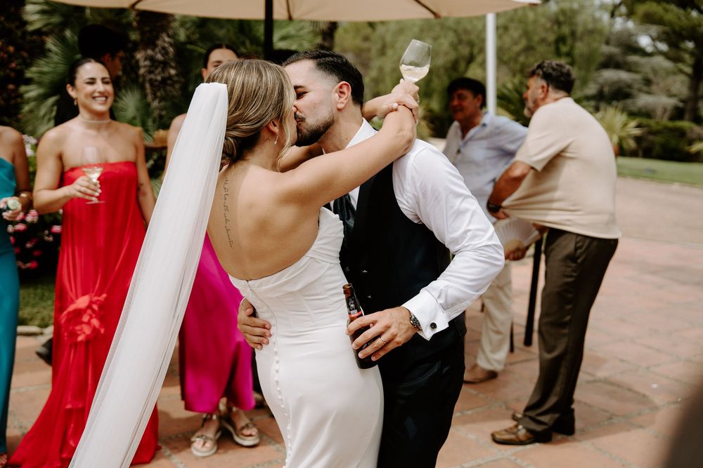 Bride and groom share a kiss during outdoor wedding reception while guests celebrate in background.