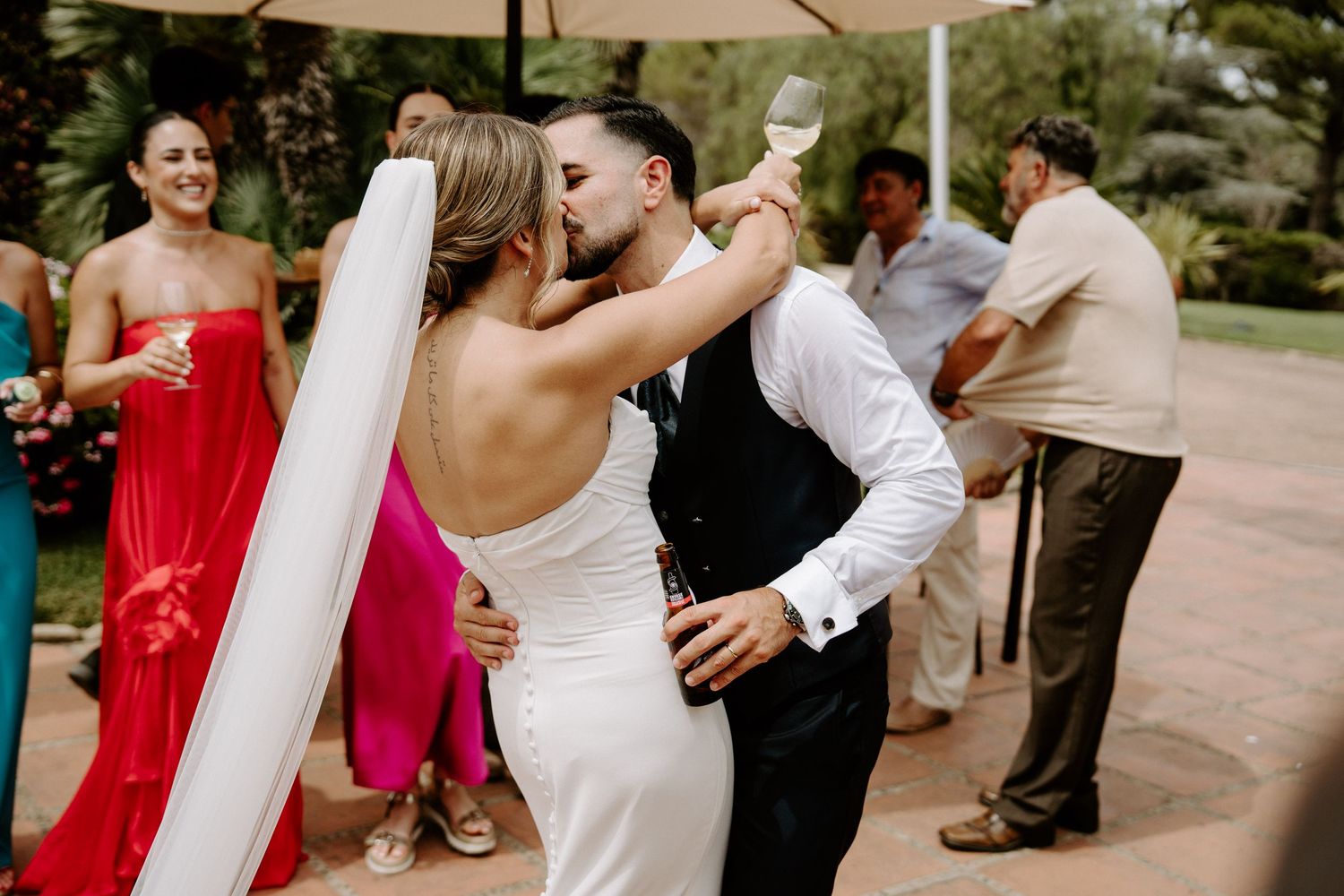 Bride and groom share a kiss during outdoor wedding reception while guests celebrate in background.