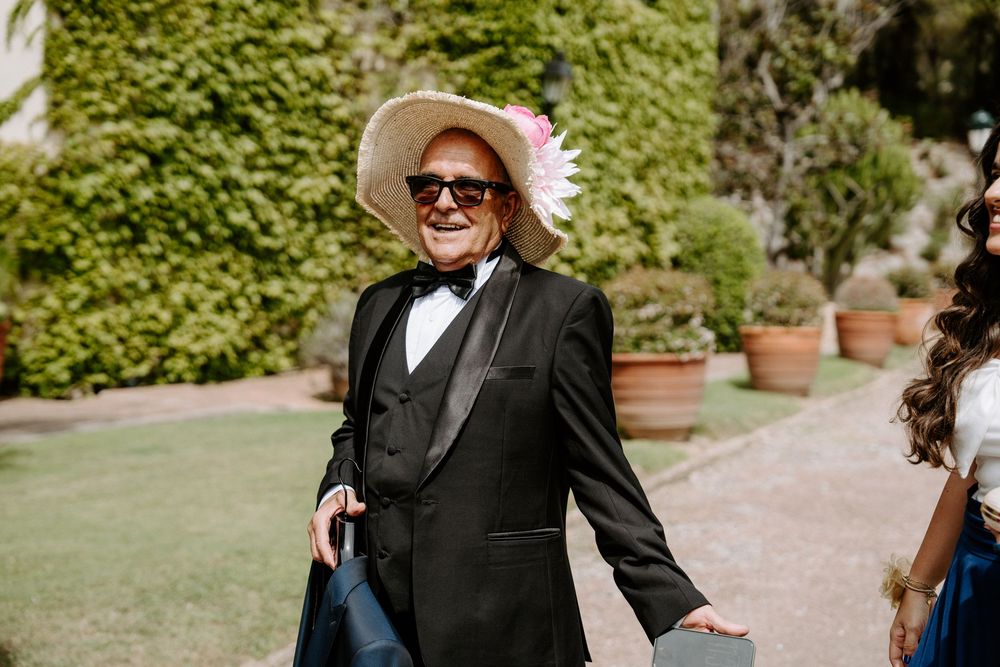 Elderly man in black tuxedo and straw hat with pink flower, smiling outdoors at a garden wedding event.
