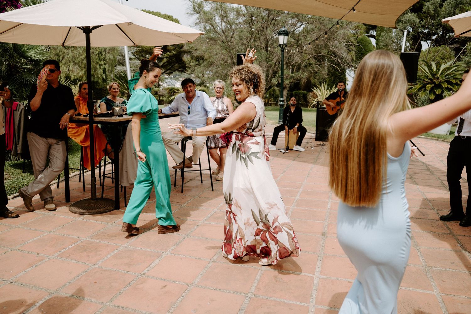 Wedding guests dancing outdoors on terrace with live band performing under umbrellas at sunny reception.