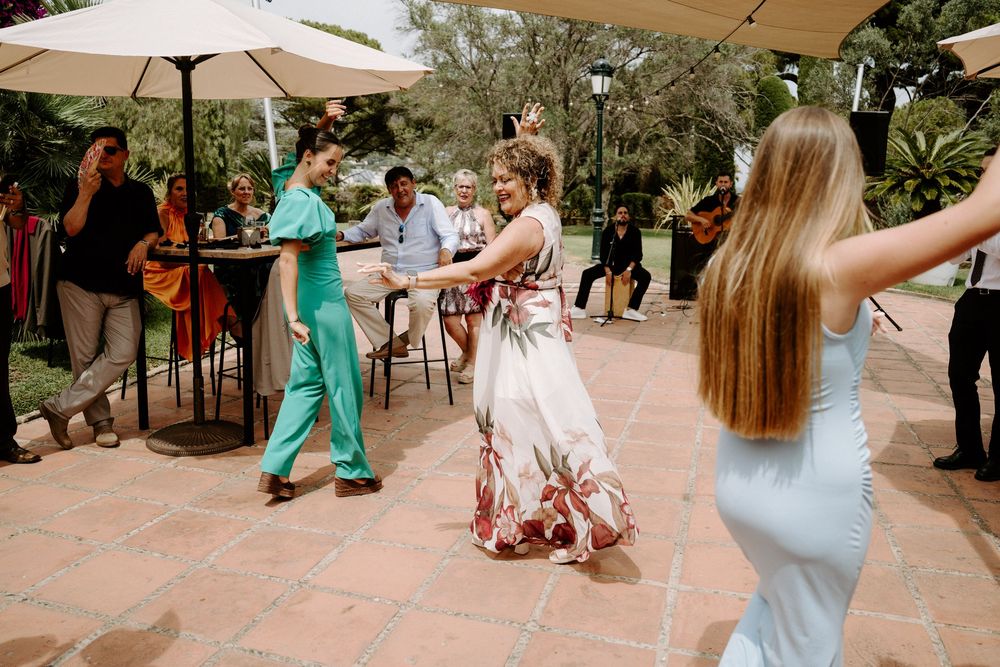 Wedding guests dancing outdoors on terrace with live band performing under umbrellas at sunny reception.