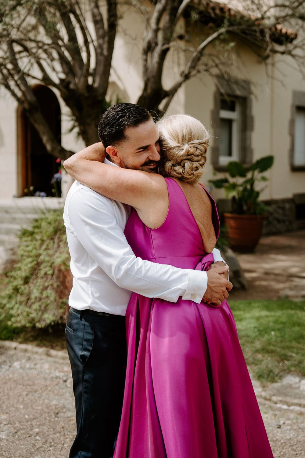 A man in a white shirt embraces a woman in a vibrant pink gown outdoors near a rustic building.