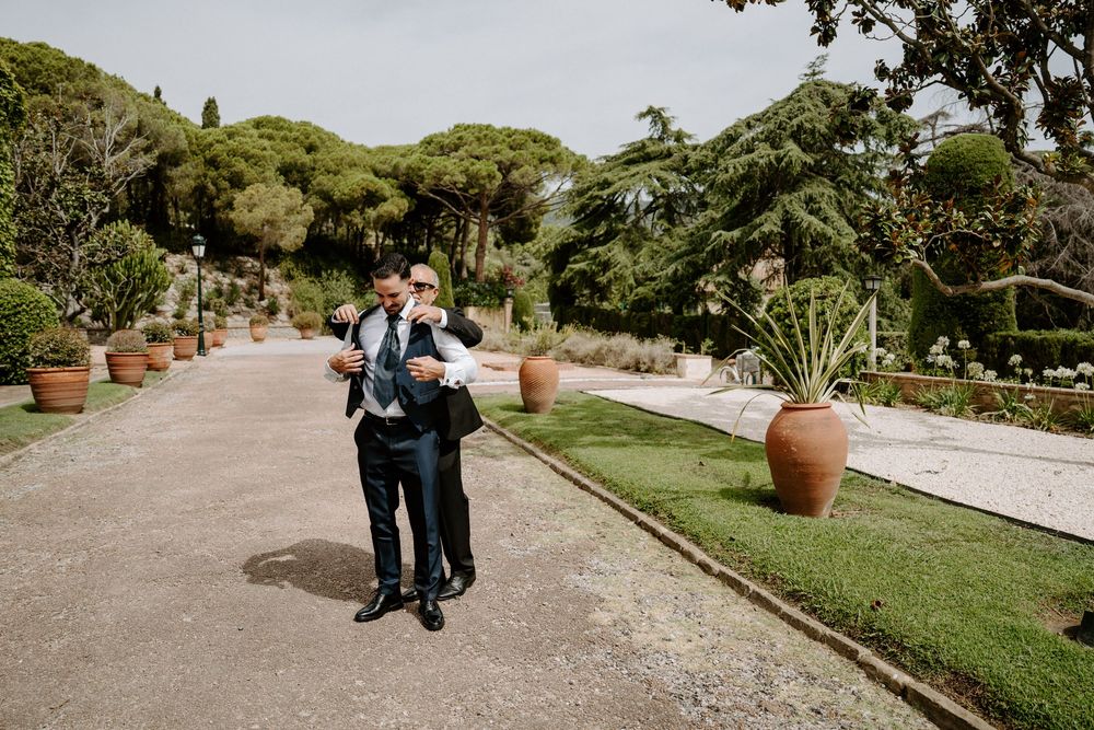 Two grooms in suits walk together through an elegant Italian garden with terracotta pots and lush greenery.