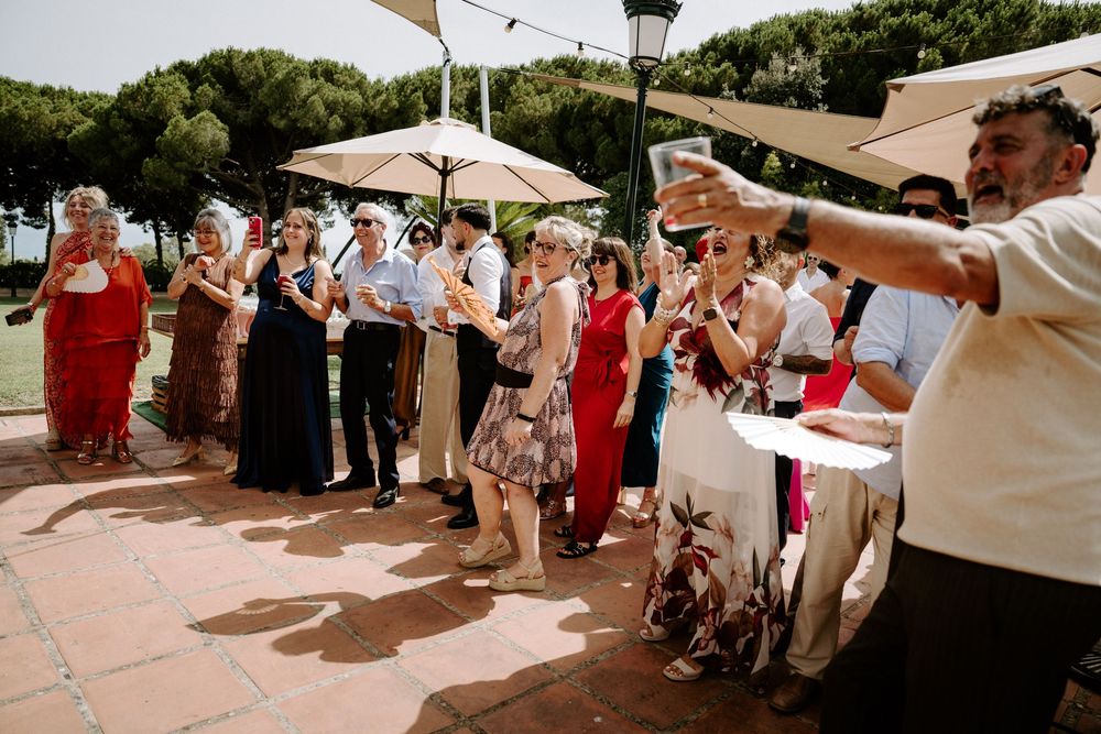 Wedding guests gathered outdoors under umbrellas participating in a lively celebration activity together.