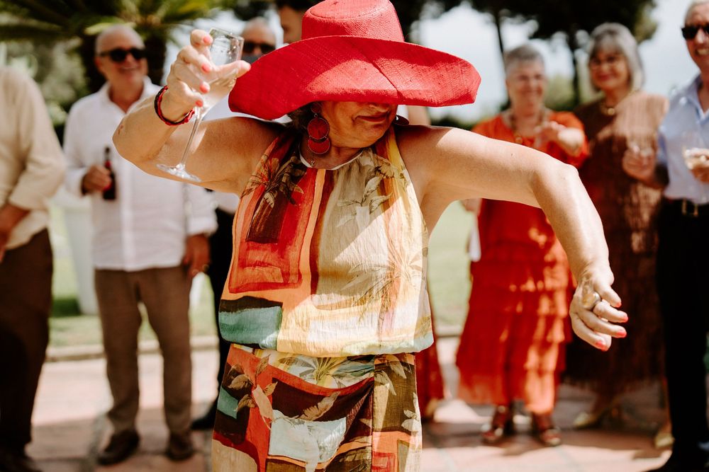 Woman in red hat and colorful dress dancing at outdoor wedding celebration.