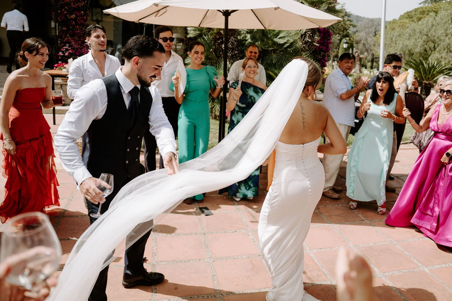 Bride and groom dance joyfully outdoors as wedding veil flows in the breeze among celebrating guests.
