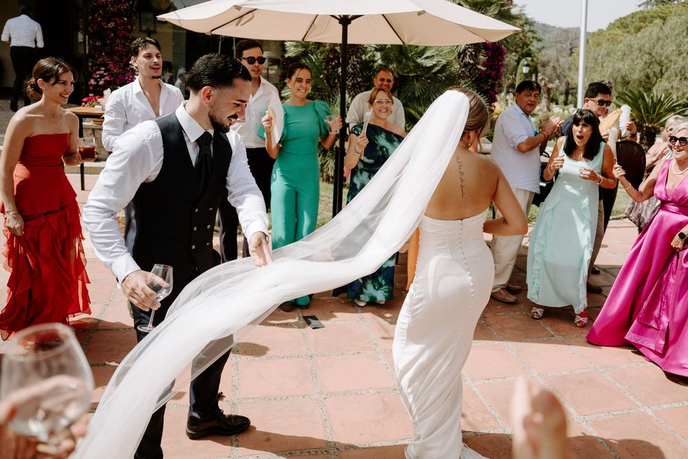 Bride and groom dance joyfully outdoors as wedding veil flows in the breeze among celebrating guests.
