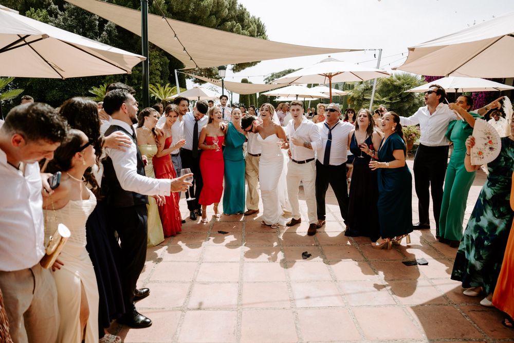 Wedding couple shares first dance surrounded by cheering guests at sunny outdoor reception venue.