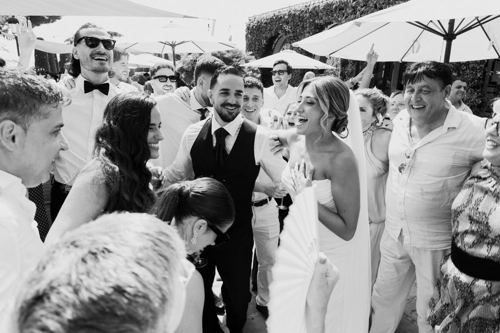 Black and white photo of bride and groom laughing with wedding guests at outdoor reception.