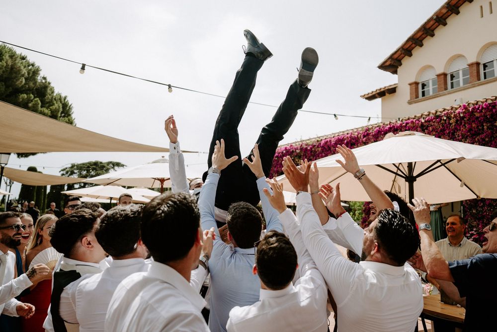 Groomsmen tossing groom into the air during lively outdoor wedding celebration under string lights.