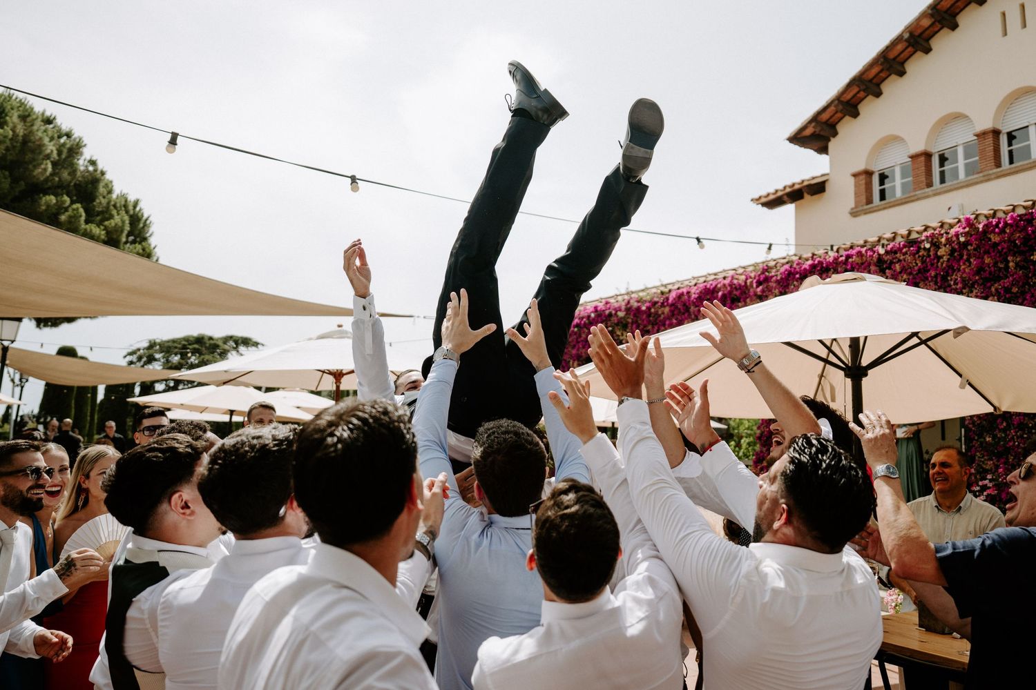 Groomsmen tossing groom into the air during lively outdoor wedding celebration under string lights.
